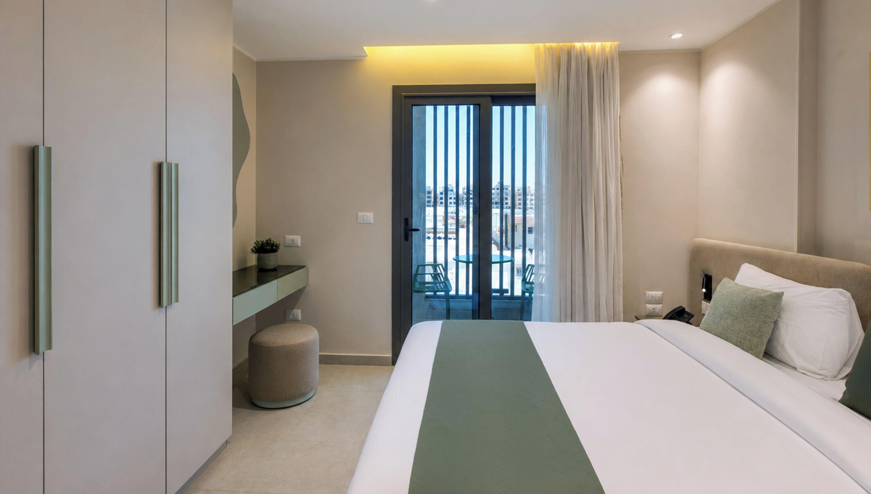 A hotel room with a bed, a nightstand, a small table, a chair, and a view of the city through a sliding door.