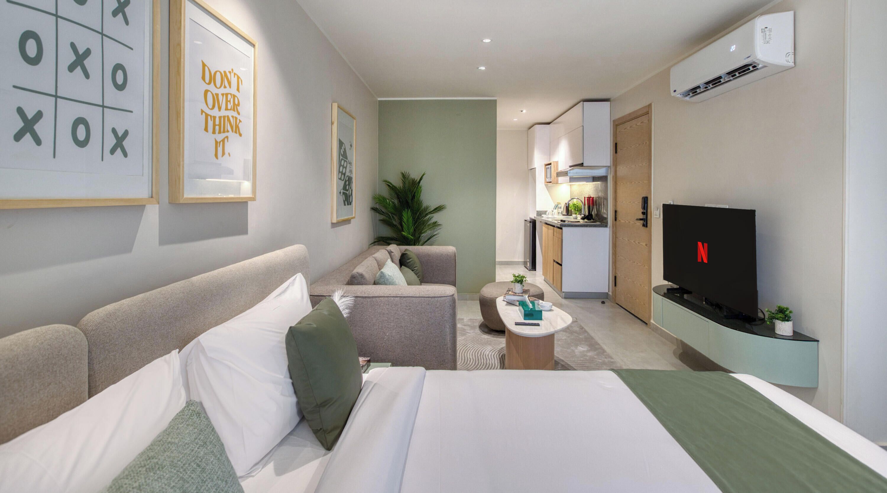 A hotel room with a bed, a sofa, a small coffee table, a TV, and a kitchenette in the background.