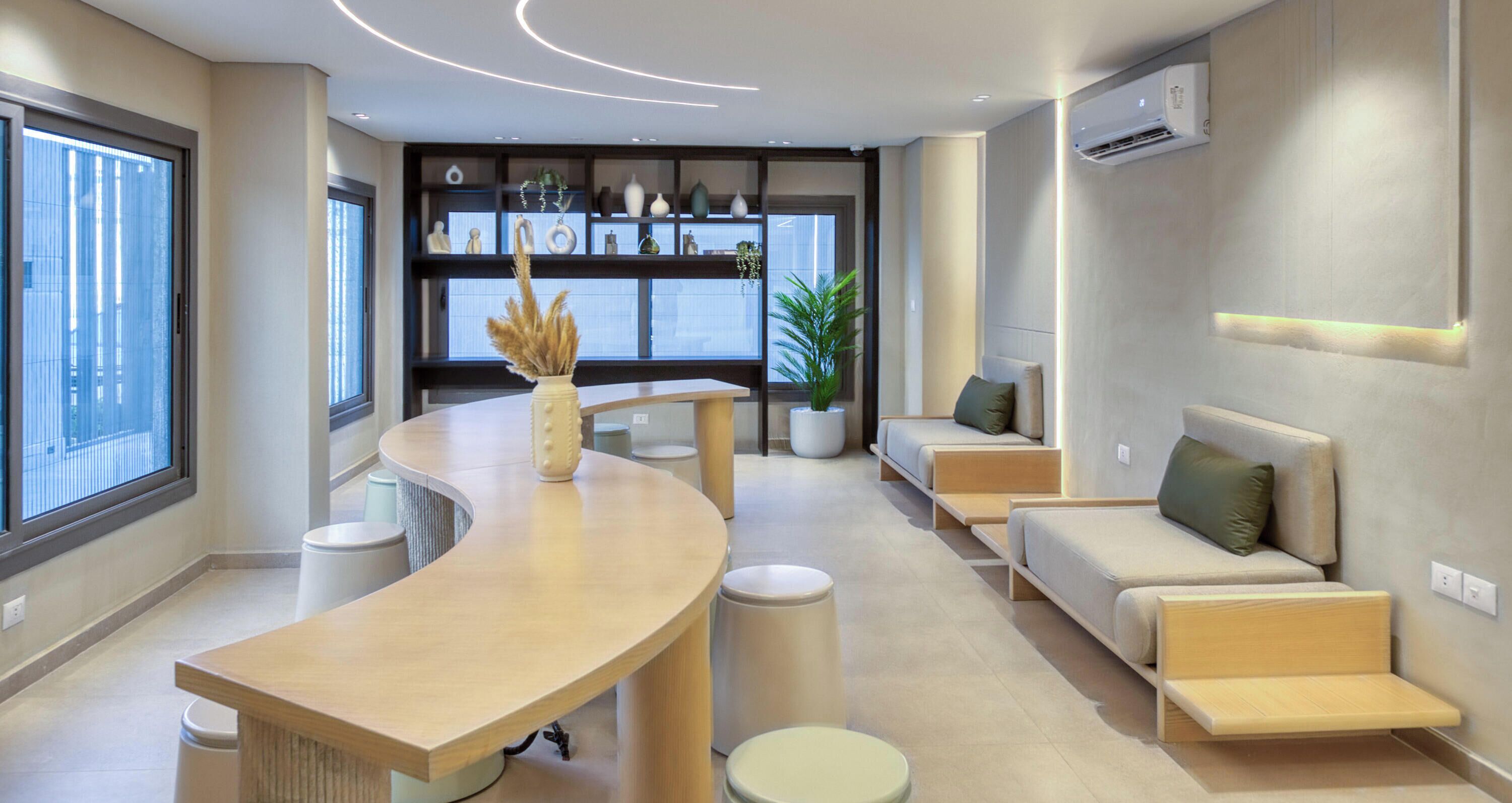 A modern interior with a curved wooden counter, seating area with cushions, a potted plant, and a built-in shelf with decorative items.
