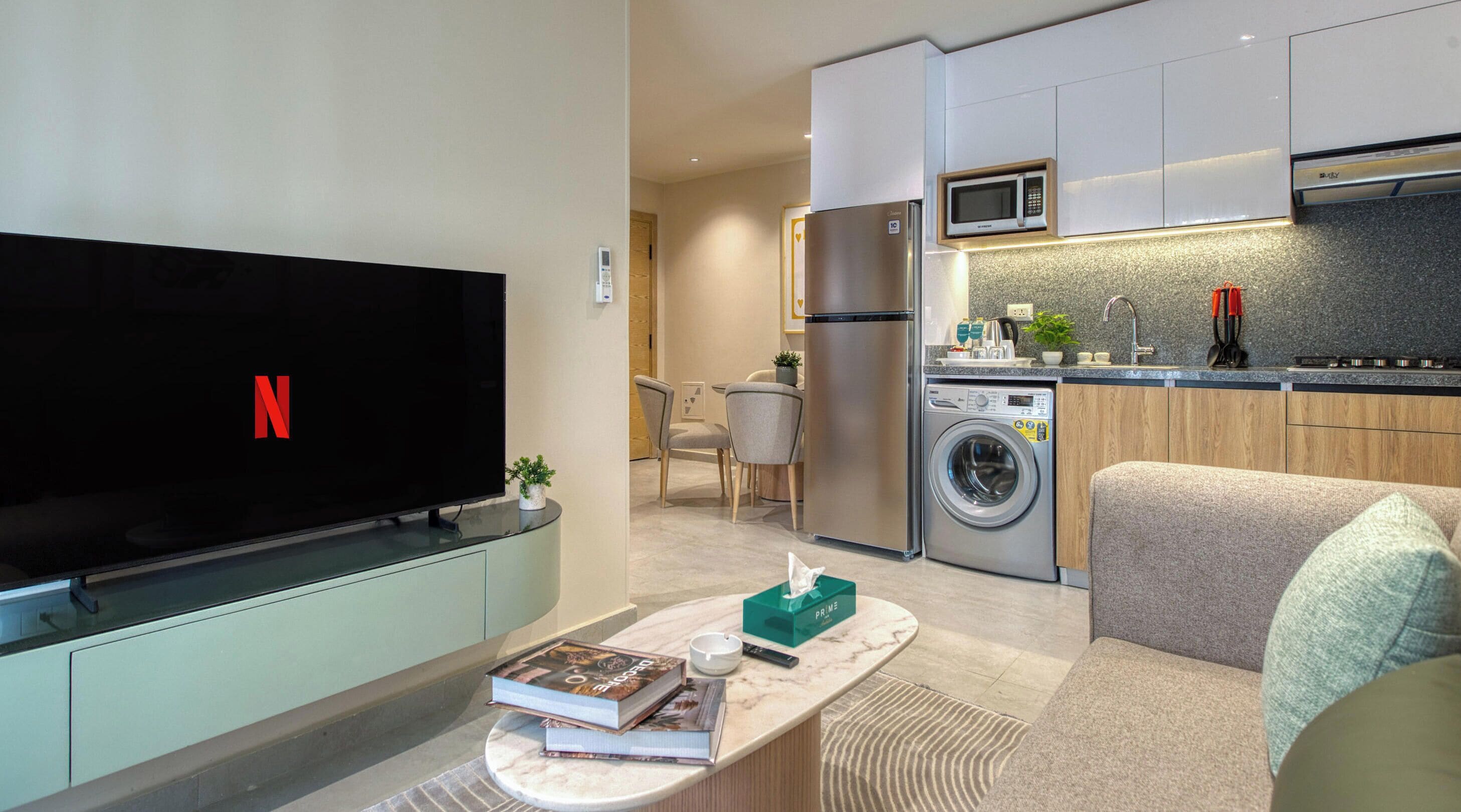 Una sala de estar moderna con un televisor de pantalla plana, un sofá, una mesa de centro con libros y un control remoto, una lavadora y una zona de cocina con microondas y fregadero.