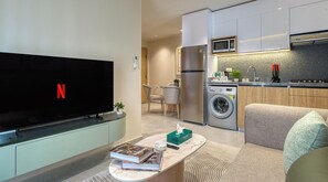 A modern living room with a flat-screen TV, a sofa, a coffee table with books and a remote, a washing machine, and a kitchen area with a microwave and sink.