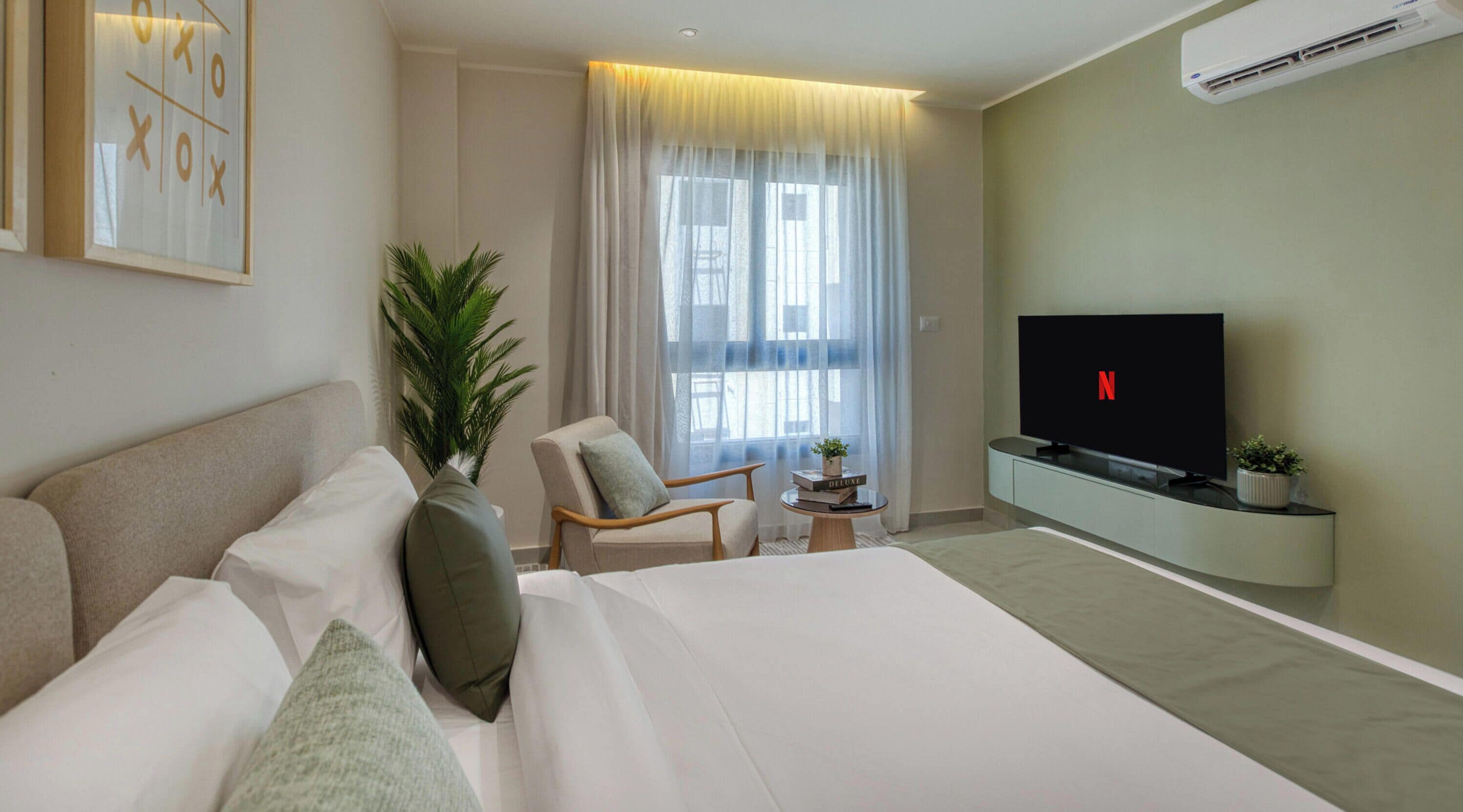 A modern hotel room with a large bed, a seating area with a chair and a small table, a television, and a potted plant.