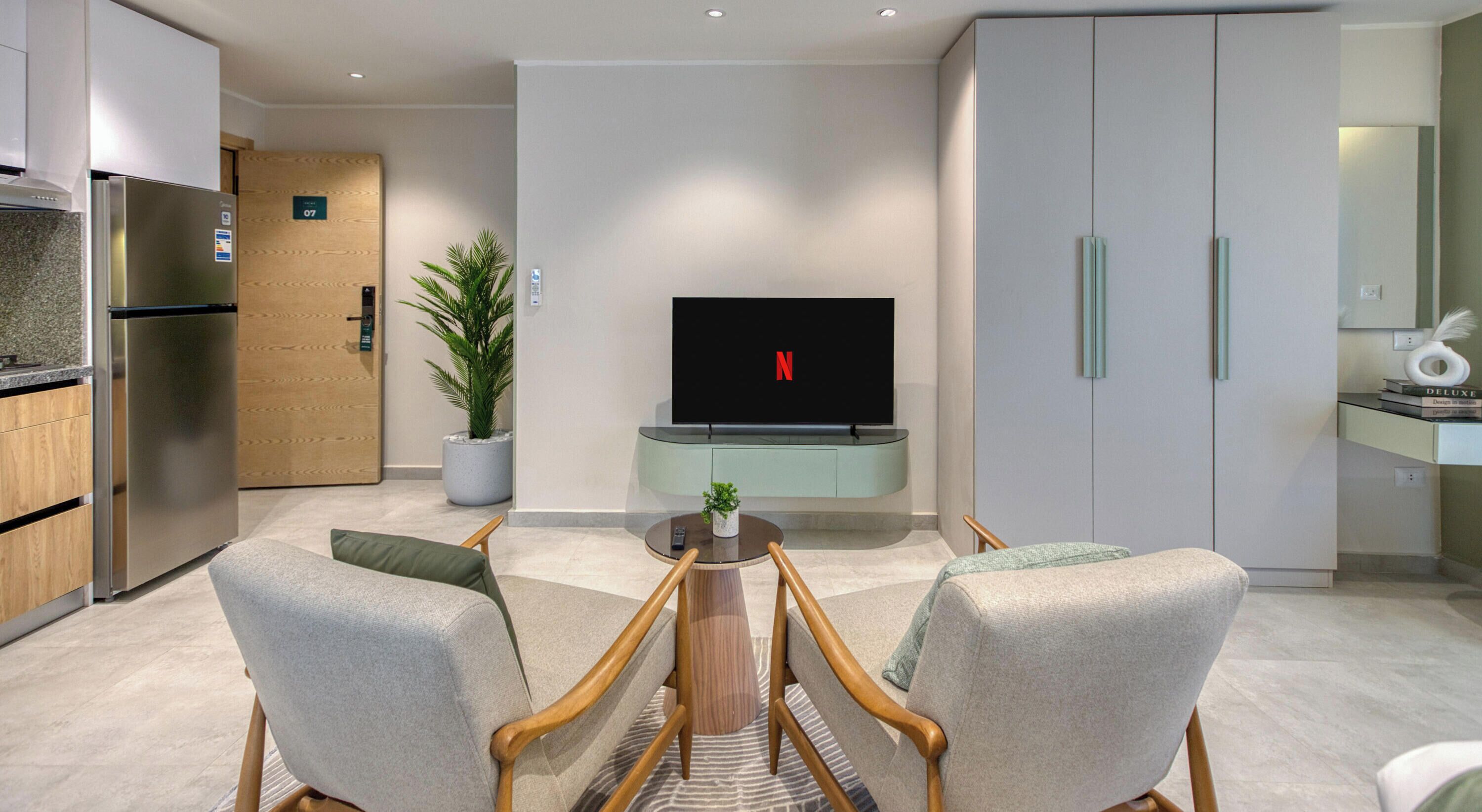 A modern living room with a flat-screen TV, a dining table with chairs, and a kitchen area with a refrigerator.