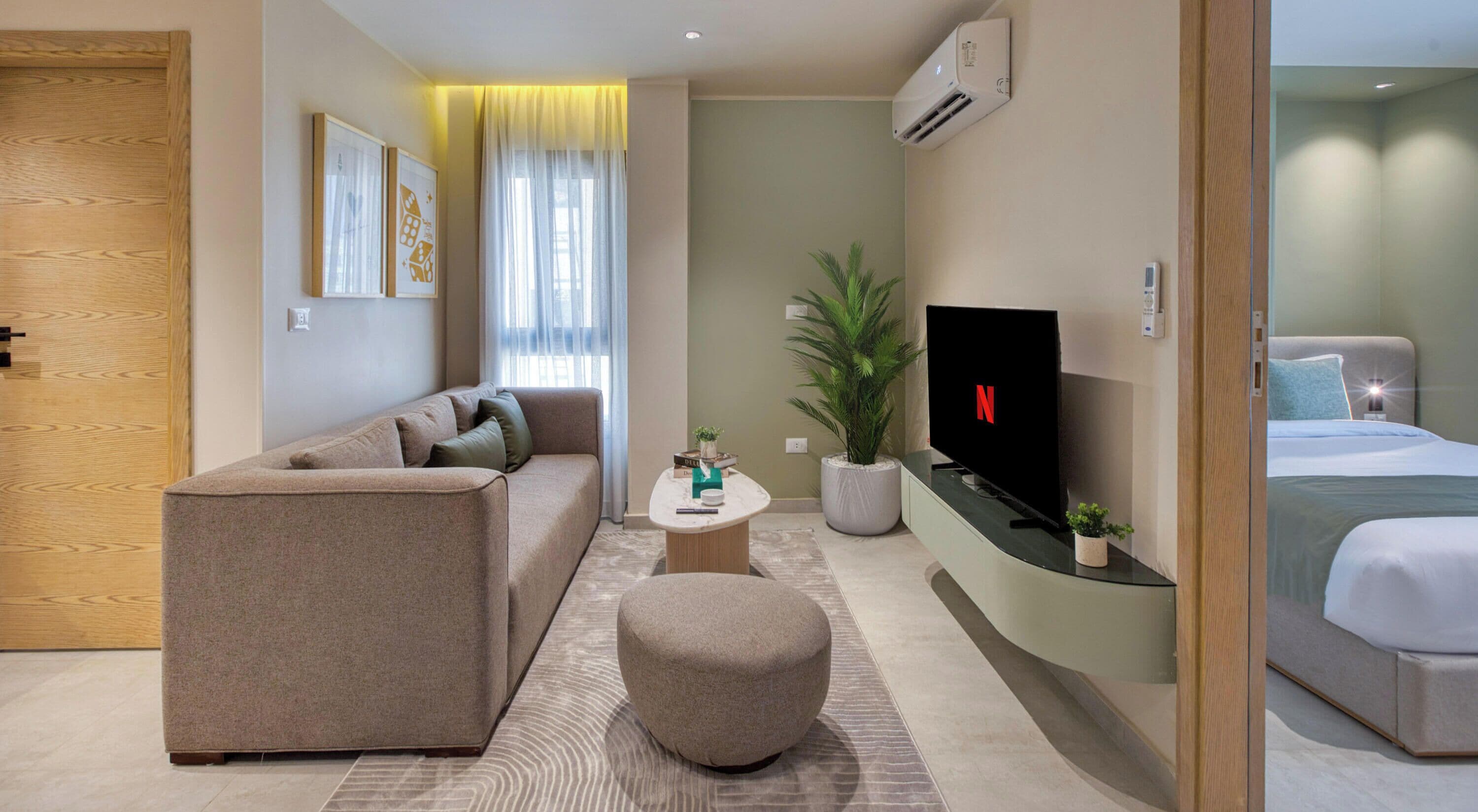 Quarto de hotel moderno com sofá, mesa de centro, televisão, cama e planta em vaso.