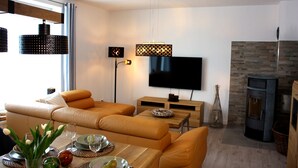 A modern living room with a sofa set, a flat-screen TV, a fireplace, and a dining area with a table set for two. - Semi-detached house - 4 bedrooms for families and friends (Jesendorf)