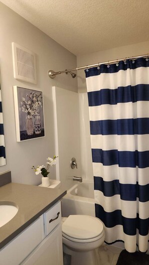 A bathroom with a white toilet, a bathtub, a shower curtain, a vent, and a vase with flowers on the counter. - Pet-Friendly Faribault Home • Near Shattuck-St. Mary's (Faribault)