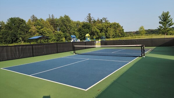 An outdoor tennis court with a net, surrounded by a fence and trees.