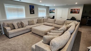 A living room with a large sectional sofa, a coffee table, and a fireplace. There are two windows with blinds, a door, and a kitchen area in the background.