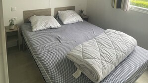 A single bed with a patterned bedspread, a bedside table with a lamp, and a window with curtains. - Mobile Home, at a Campground ⭐️⭐️⭐️⭐️⭐️ 3 km from the ocean (Saint-Arnoult)