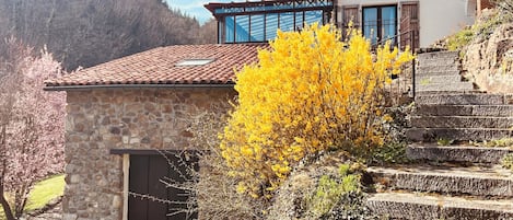 Un edificio de piedra con techo de tejas y puerta de madera, rodeado de una escalera de piedra y un árbol floreciente de color amarillo.