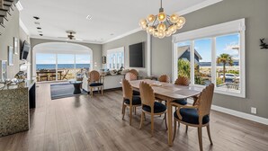 A spacious dining area with a view of the ocean, a large chandelier, and a modern kitchen in the background. - Ocean Front, Updated 4 Bedroom, 3.5 Bath (St. Simons Island)