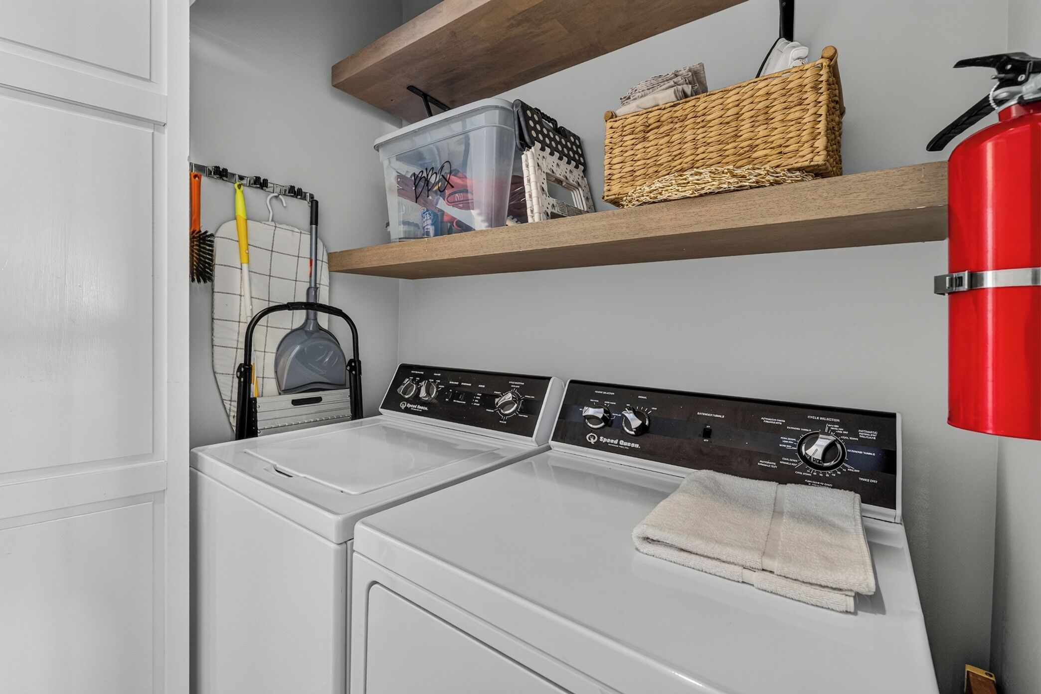 Un espace buanderie comprenant un lave-linge blanc, un sèche-linge et une étagère avec des paniers et un extincteur.