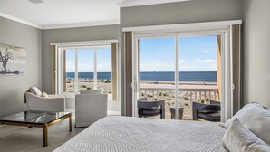 A bedroom with a large bed, a glass coffee table, two chairs, and a view of the beach through open sliding doors. - Ocean Front, Updated 4 Bedroom, 3.5 Bath (St. Simons Island)