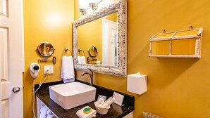 Un baño con lavamanos blanco, un espejo grande, un secador de pelo, un perchero para toallas y un jabonera.