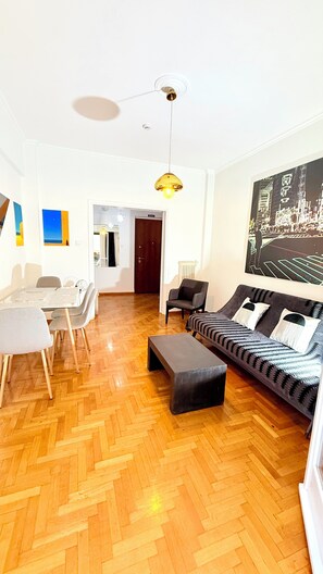 Ein modernes Wohnzimmer mit Holzdielen im Fischgrätmuster, einer Couch, einem Esstisch mit Stühlen und einer Pendelleuchte.