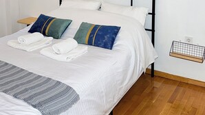Cama bem arrumada com lençóis brancos e manta de tricô cinza, ar condicionado fixado na parede e um painel de macramé.