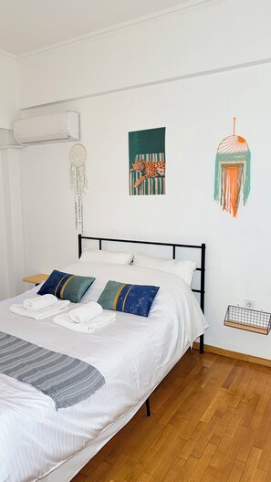 Ein ordentlich bezogenes Bett mit weißen Leinen und einer grauen Strickdecke, eine Wand-Klimaanlage und ein Wandbehang aus Makramee.