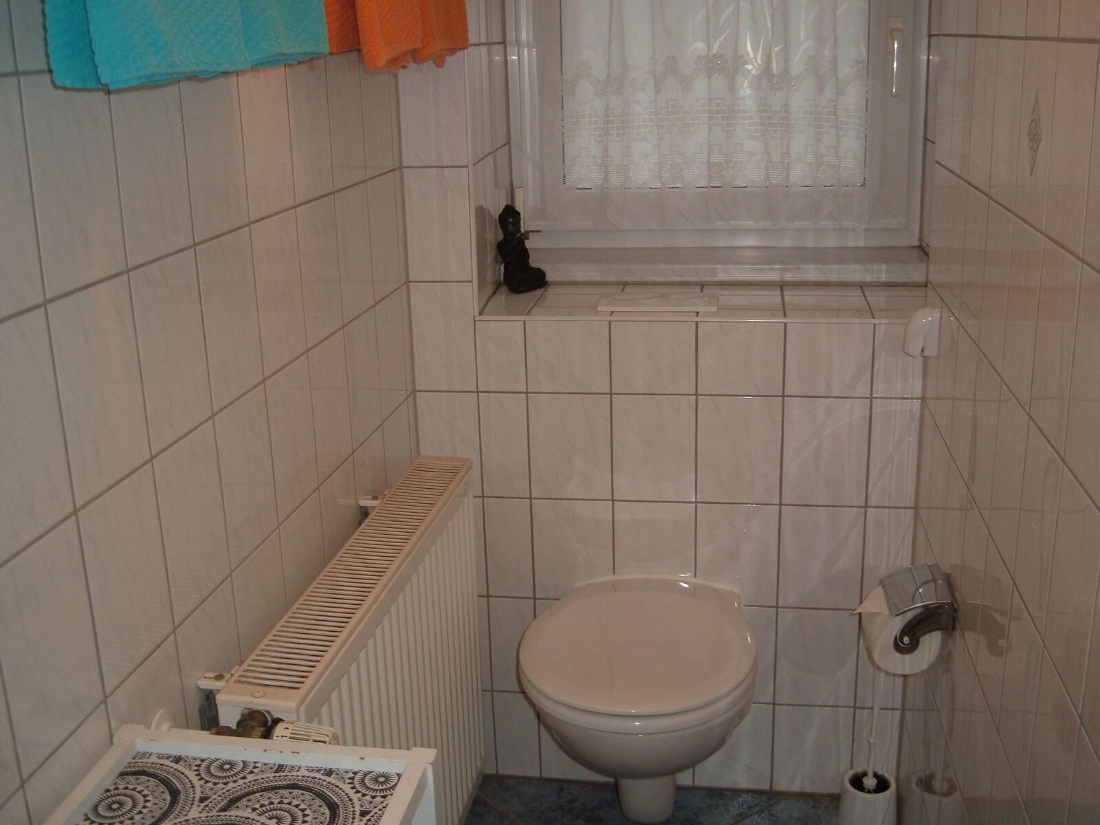 A small, tiled bathroom with a toilet, a radiator, and a window with a curtain.