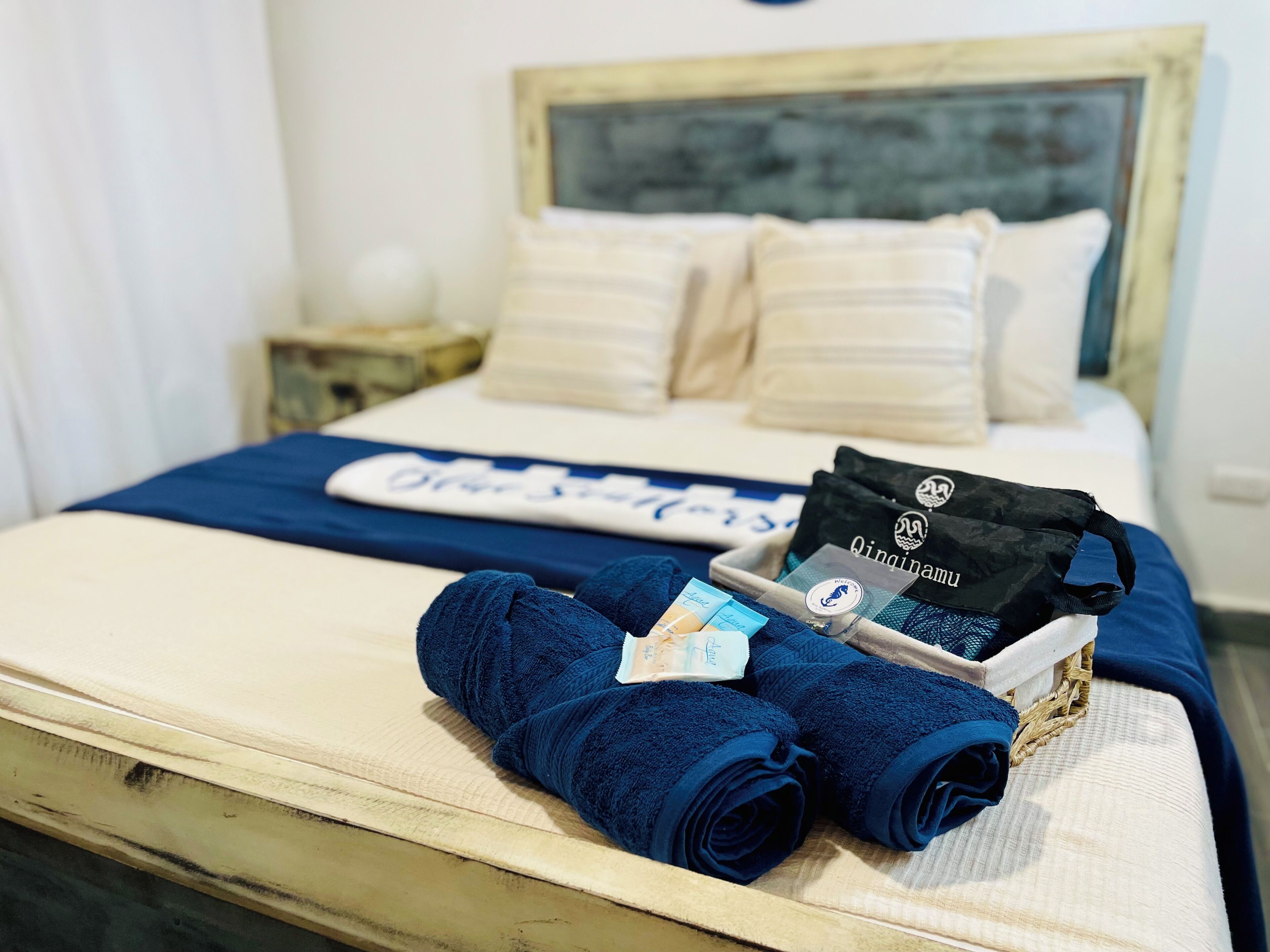 Un lit bien fait, recouvert d’une couverture bleue, une pile de serviettes pliées, un sac noir et un plateau contenant des articles de toilette.