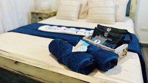 Una cama bien hecha con una manta azul, un juego de toallas dobladas, un bolso negro y una bandeja con artículos de aseo.