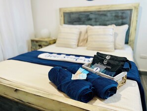 Ein ordentlich bezogenes Bett mit blauer Decke, ein Satz gefalteter Handtücher, eine schwarze Tasche und ein Tablett mit Pflegeprodukten.