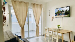 Una sala de estar con un sofá, una mesa de comedor con sillas, un televisor montado en la pared y un balcón con cortinas.