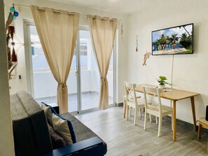 Ein Wohnzimmer mit Sofa, einem Esstisch mit Stühlen, einem an der Wand montierten Fernseher und einem Balkon mit Vorhängen.