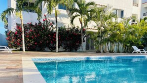 Una piscina con agua azul cristalina, rodeada de palmeras y flores rojas, con sillas de descanso blancas y un edificio blanco al fondo.
