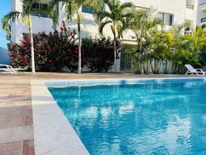 Ein Swimmingpool mit klarem, blauem Wasser, umgeben von Palmen und roten Blüten, mit weißen Liegestühlen und einem Gebäude im Hintergrund.