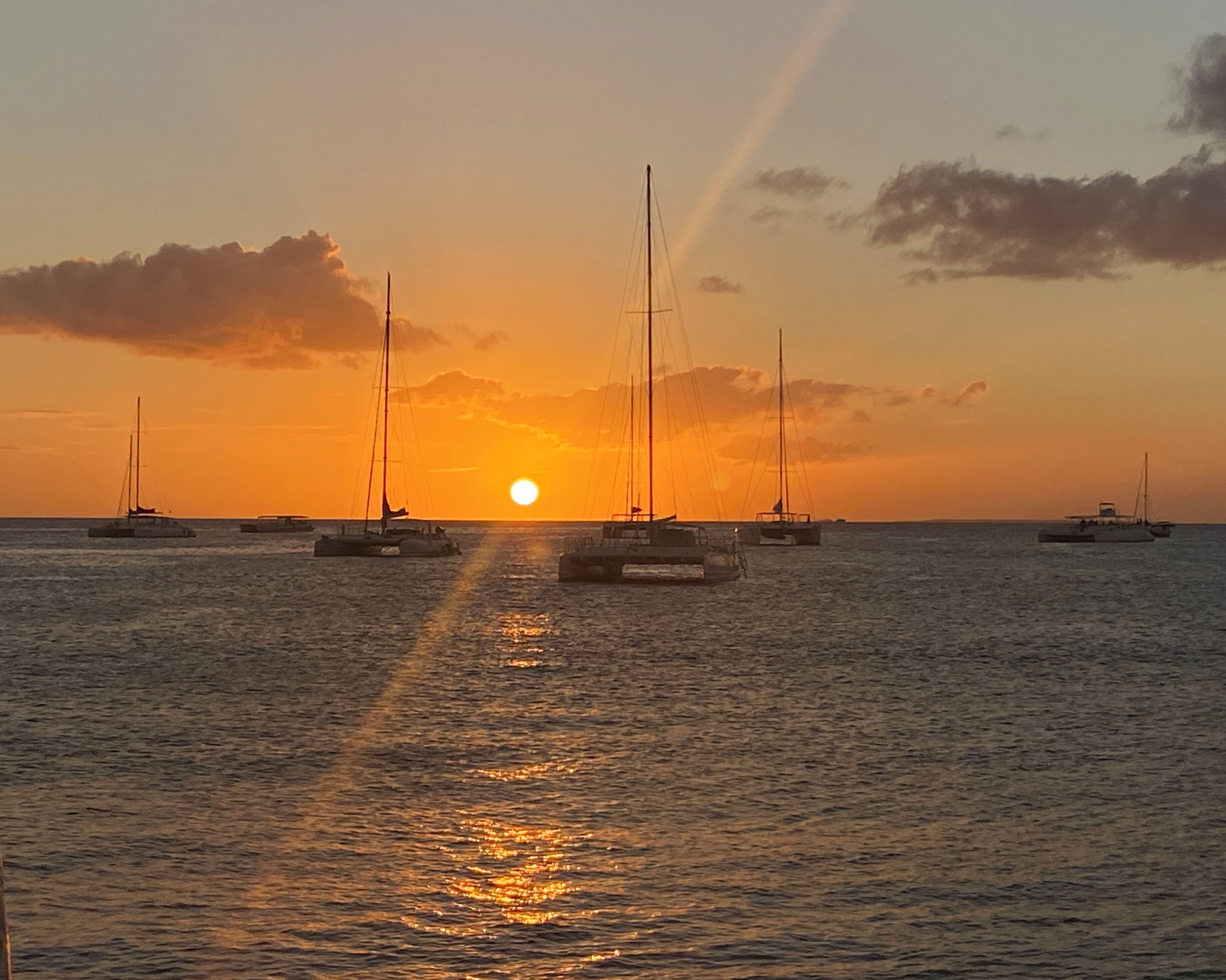 A sunset over calm waters with several sailboats anchored.