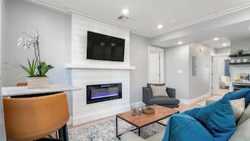 A modern living room with a fireplace, a flat-screen TV mounted above it, a sofa, a coffee table, and a dining area with a table and chairs.