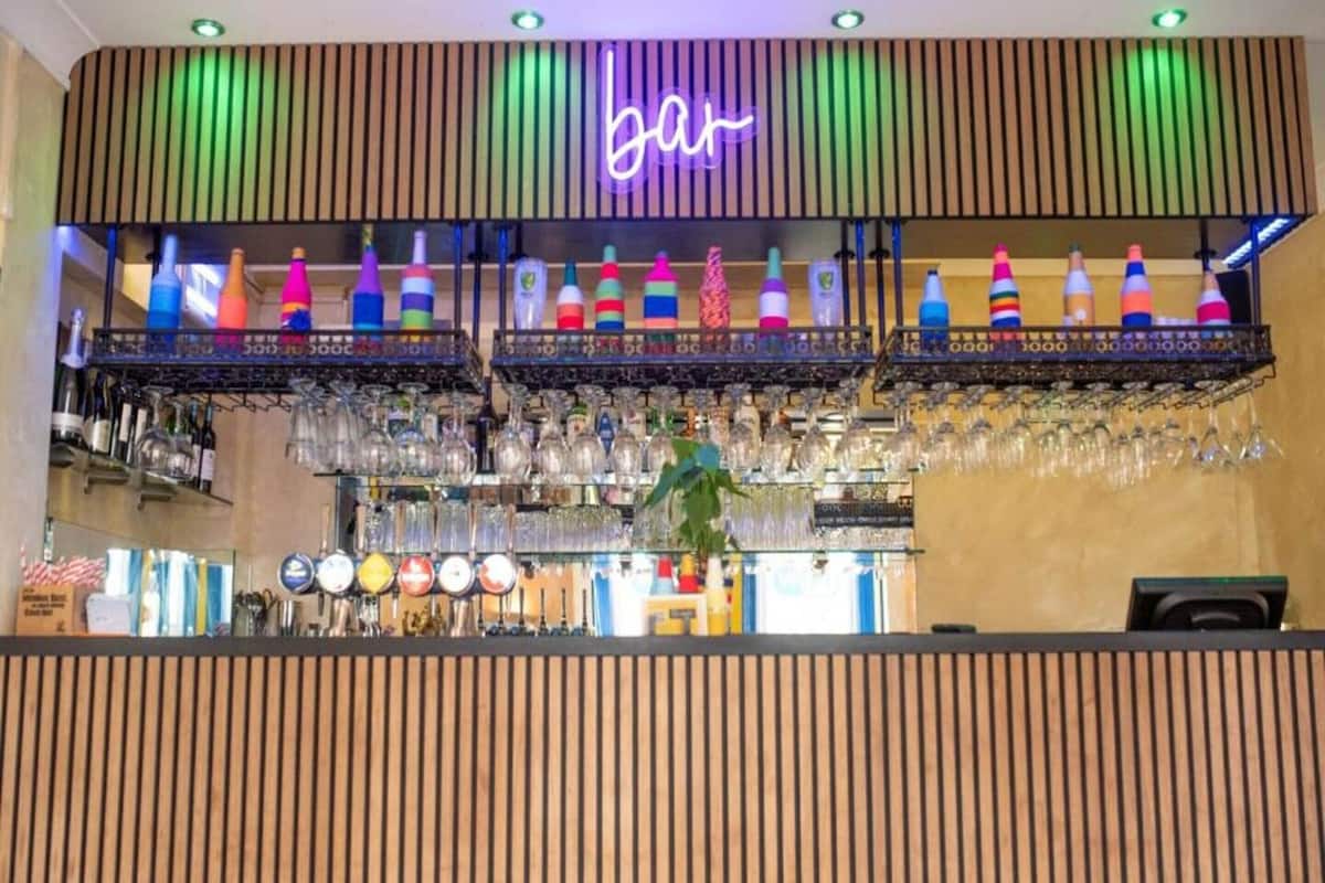 Zona de bar con un letrero de neón, estantes con botellas de colores y diversos vasos.