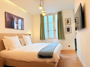 Ein Hotelzimmer mit Bett, Fenster mit Vorhängen, einem an der Wand befestigten Fernseher und Kunstwerken.