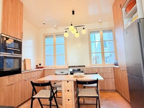 Eine moderne Küche mit Holzschränken, einem eingebauten Backofen, einem Esstisch mit Stühlen und einem großen Fenster, das natürliches Licht hereinlässt.