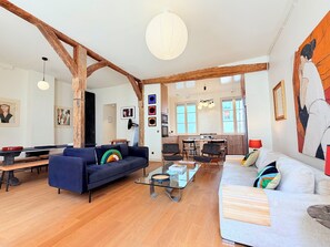 Ein geräumiges Wohnzimmer mit einer großen Couch, einer kleineren Couch, einem Couchtisch und einem Essbereich mit Stühlen. Im Hintergrund ist eine Küche mit Esstisch und Stühlen zu sehen.