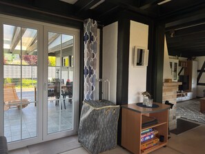 Ein modernes Interieur mit einer Glastür, die auf eine Terrassengrundstück führt, einem gemusterten Hocker und einem Holztisch mit Büchern.
