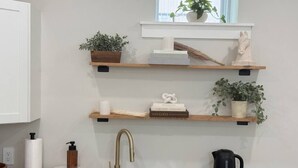 Une cuisine dotée d’un robinet moderne, d’étagères en bois ornées de plantes et de livres, d’une bouilloire et d’un plan de travail en marbre.