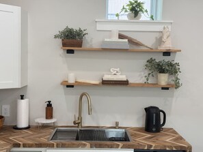 A kitchen with a modern sink, wooden shelves, and a coffee maker. - #33- Newly Constructed - Luxury Collection (Plymouth)