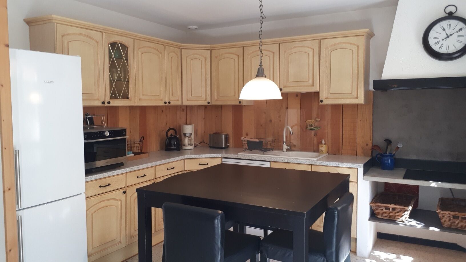 Eine Küche mit Holzausstattung, einem weißen Kühlschrank, einem schwarzen Esstisch mit Stühlen, einer Pendelleuchte und einer Wanduhr.