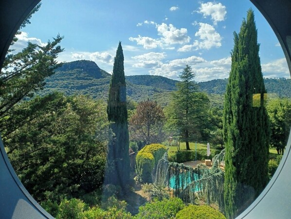 A view through a window of a lush garden with tall trees, a clear sky, and distant hills.