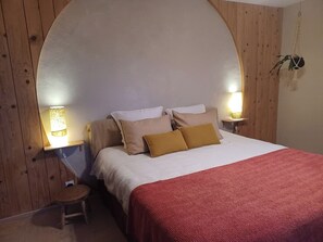 Un dormitorio con cabecera curvada, dos lámparas de noche, un taburete pequeño y una manta roja.