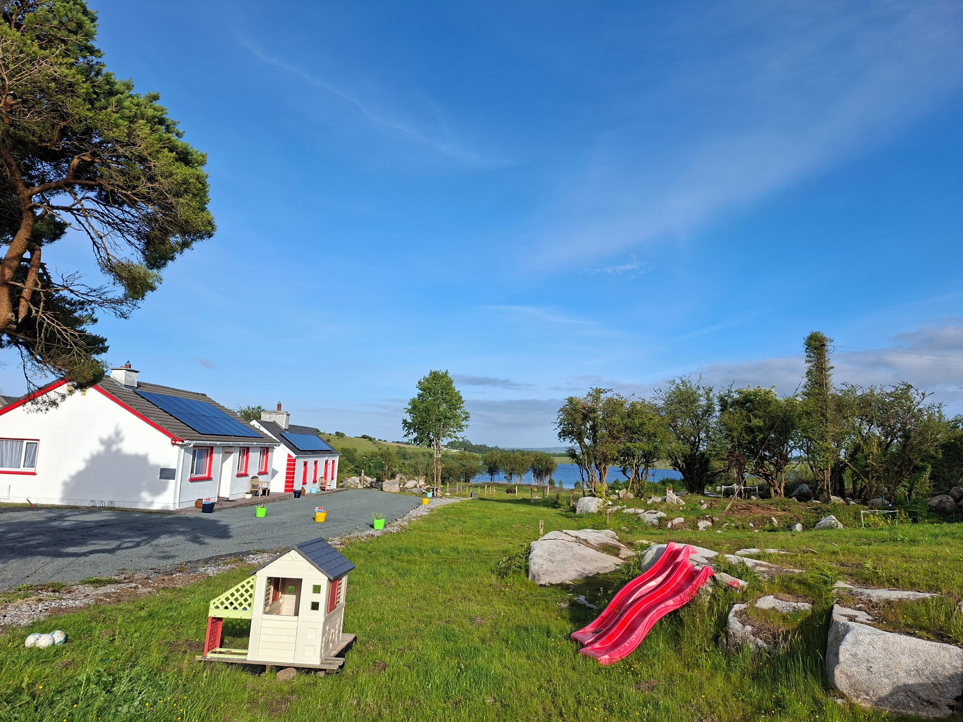 A white building with red trim, a small house, and a red slide in a grassy area with trees and a body of water in the background.