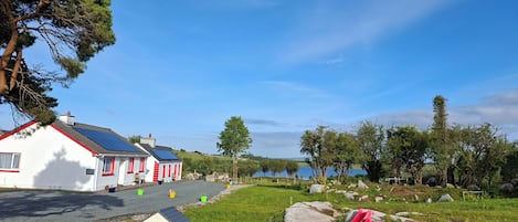 A white building with red trim, a small house, and a red slide in a grassy area with trees and a body of water in the background.