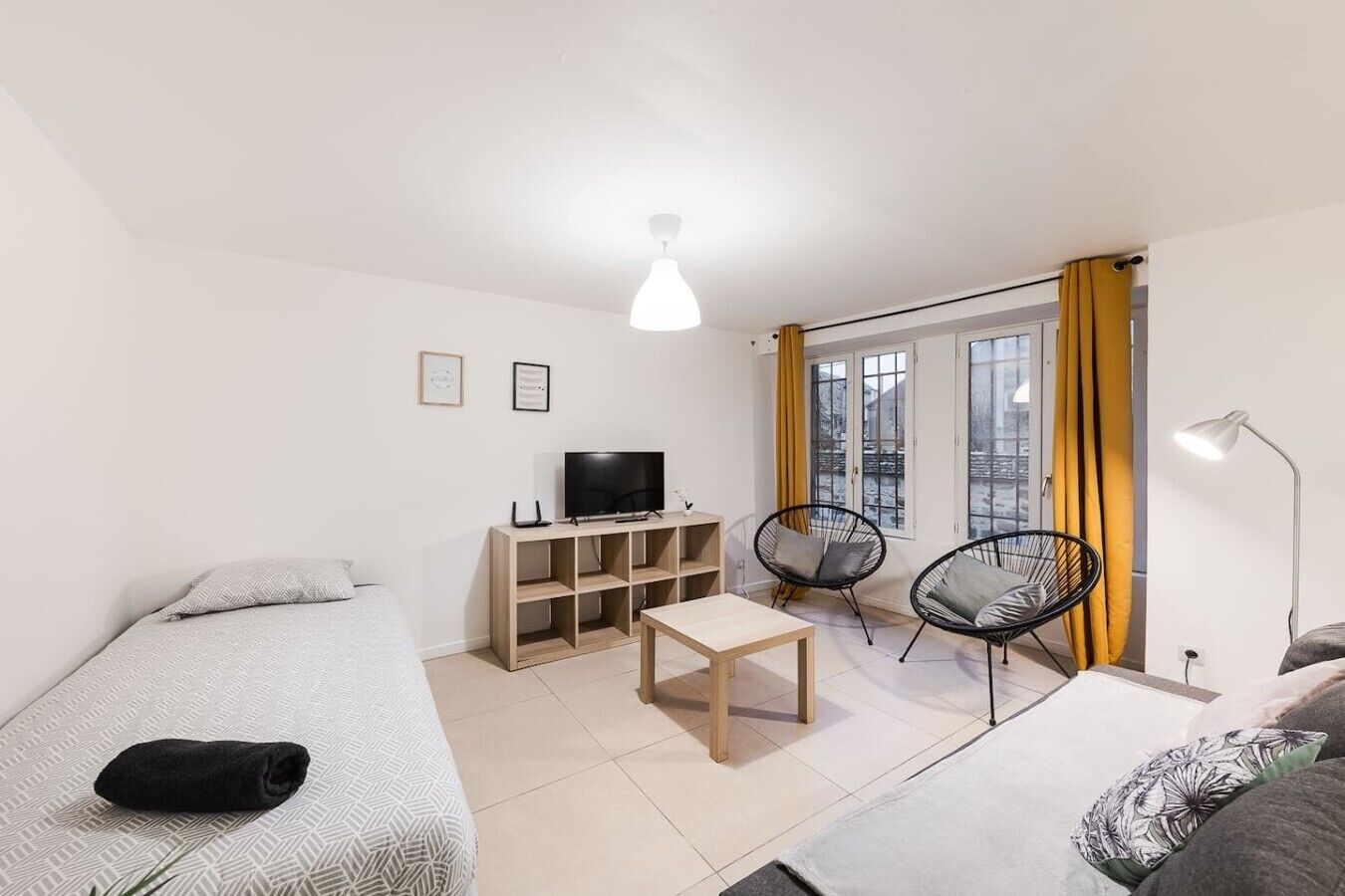 Una camera moderna e luminosa con un letto, una televisione su un mobile in legno, un tavolino, due sedie e una finestra con tende.