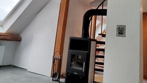 Et værelse med skråloft, indbygget træbjælke konstruktion, en sort træovn, et ovenlysvindue og en trappe til en overetage.