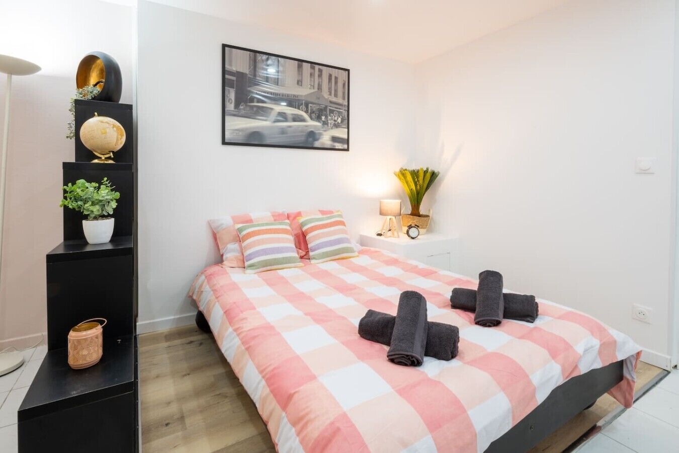 Quarto bem organizado com cama, almofadas, mesa de cabeceira com abajur e um quadro pendurado na parede.