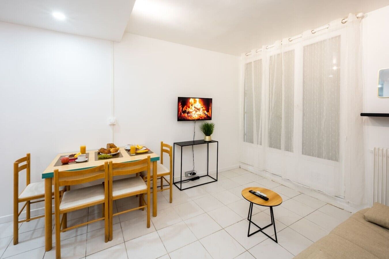 Un coin repas avec une table et des chaises, un téléviseur diffusant une image de feu de cheminée, une petite plante et une télécommande posée sur le sol.
