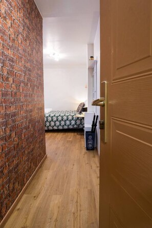 Ein Zimmer mit Backsteinwand, einem Bett mit gemustertem Überwurf, einem kleinen Tisch und einer Mülltonne.