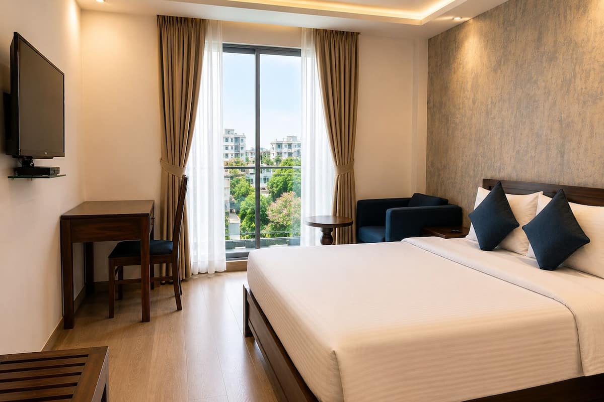 Habitación de hotel con cama, escritorio, silla, televisión y vistas a edificios y árboles.