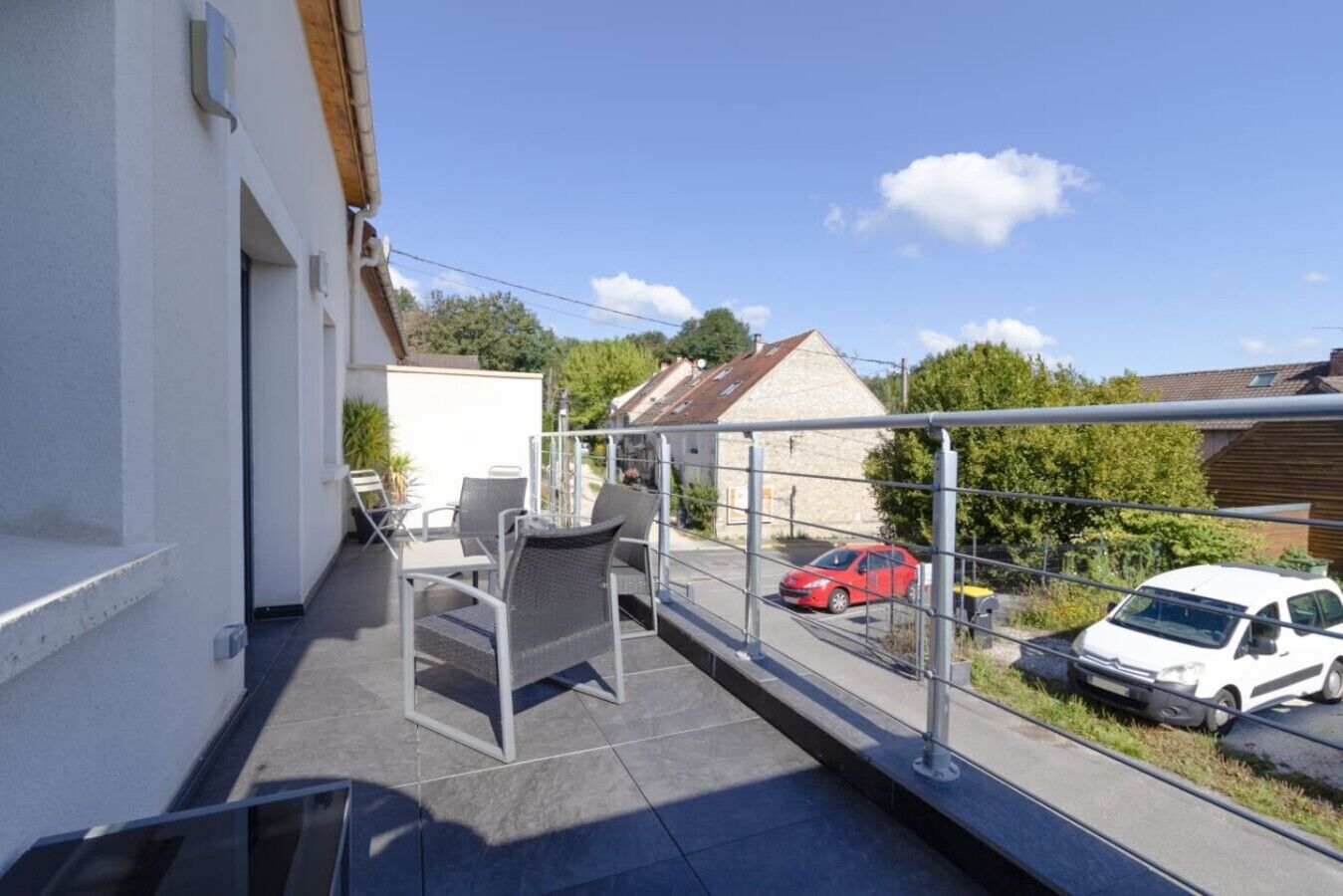 Ein Balkon mit Gartenmöbeln, Blick auf eine Straße mit parkenden Autos und ein Gebäude im Hintergrund.